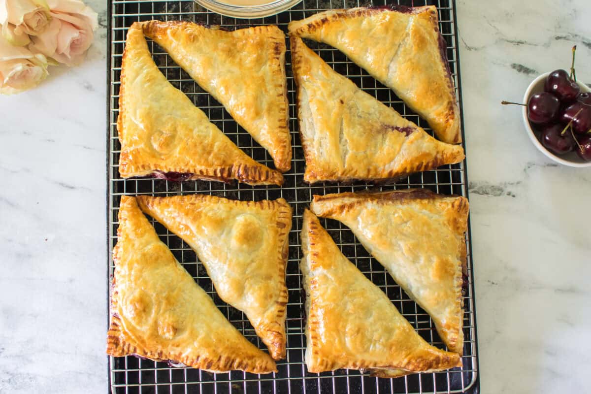 An overhead shot of eight freshly baked, triangular cherry turnovers cooling on a wire rack, featuring a deep golden-brown, puffed pastry crust with crimped edges and no glaze yet, flanked by a bowl of fresh cherries and pale pink roses.