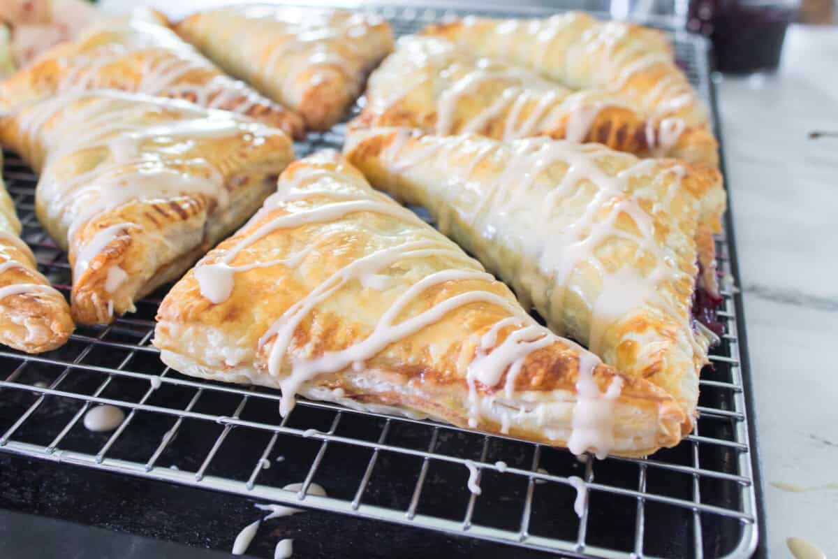A close-up shot of a flaky cherry turnover sliced in half and stacked on a wire cooling rack, revealing a thick, dark red cherry filling inside the golden puff pastry layers, drizzled with white icing glaze and garnished with fresh whole cherries.