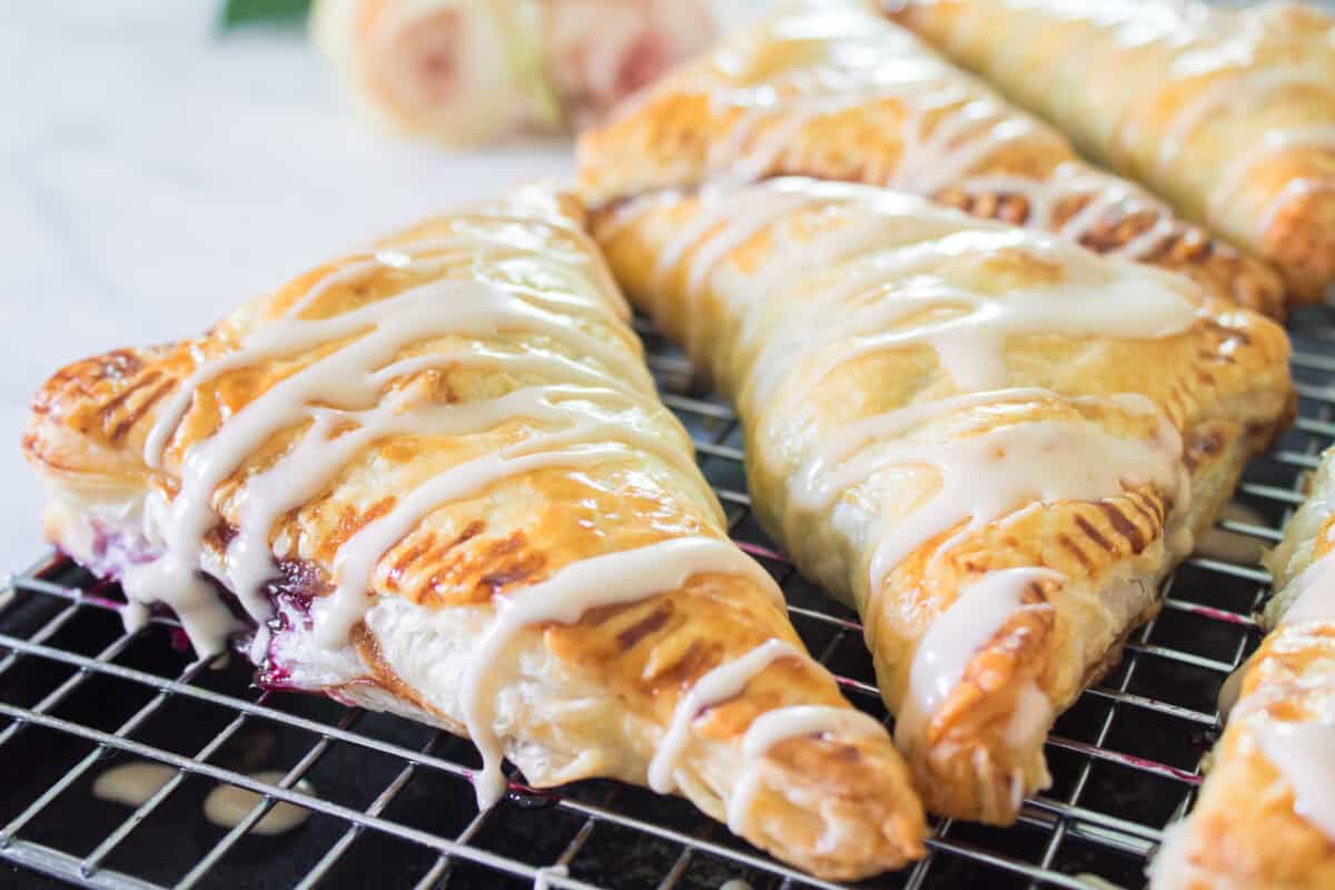 A row of golden brown, freshly baked triangular cherry turnovers cooling on a wire rack, featuring crisp fork-crimped edges and a generous drizzle of white almond glaze zigzagged across the flaky crusts.