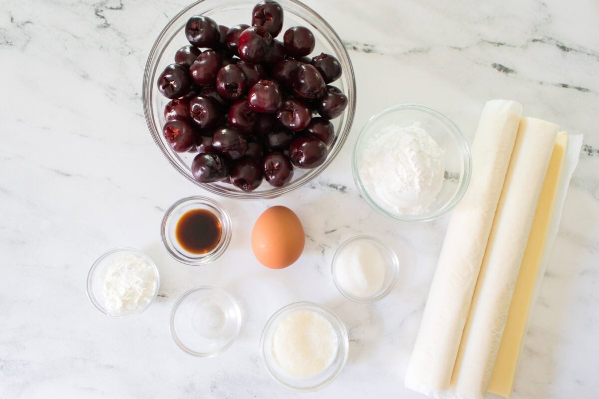 A flat lay ingredient shot on a marble countertop showing a glass bowl of pitted dark sweet cherries, rolls of puff pastry sheets, a single egg, powdered sugar, cornstarch, almond extract, heavy cream, and Purecane sweetener in small glass prep bowls.