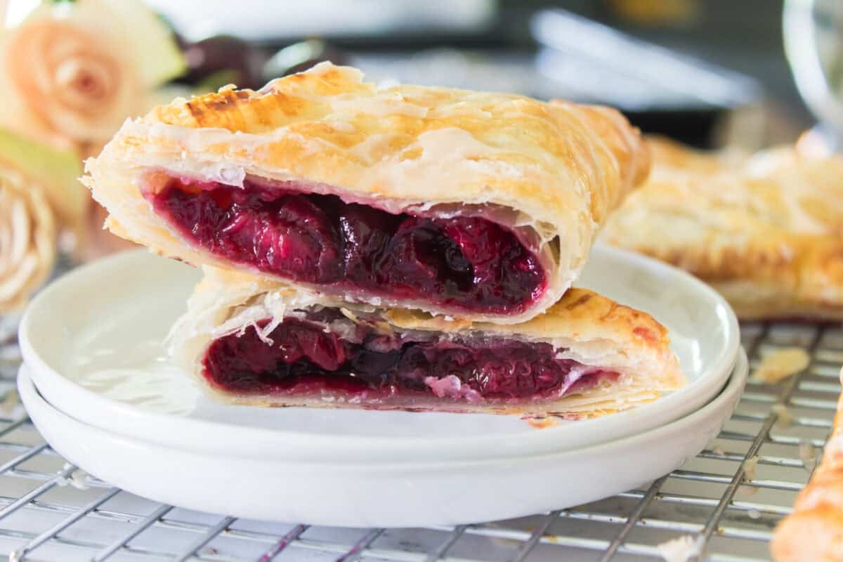 A side-angle shot of a golden cherry turnover cut in half and stacked horizontally on a small white plate, showing off the thick, jammy dark red cherry filling packed inside layers of flaky, puffed pastry.