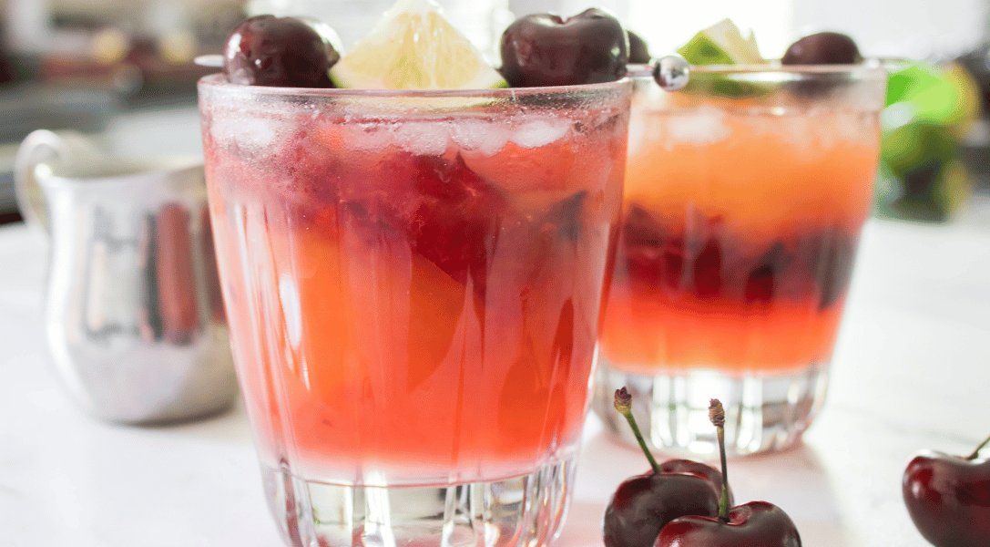2 glasses of Cherry Mango Margarita on white counter surrounded by a few dark sweet cherries