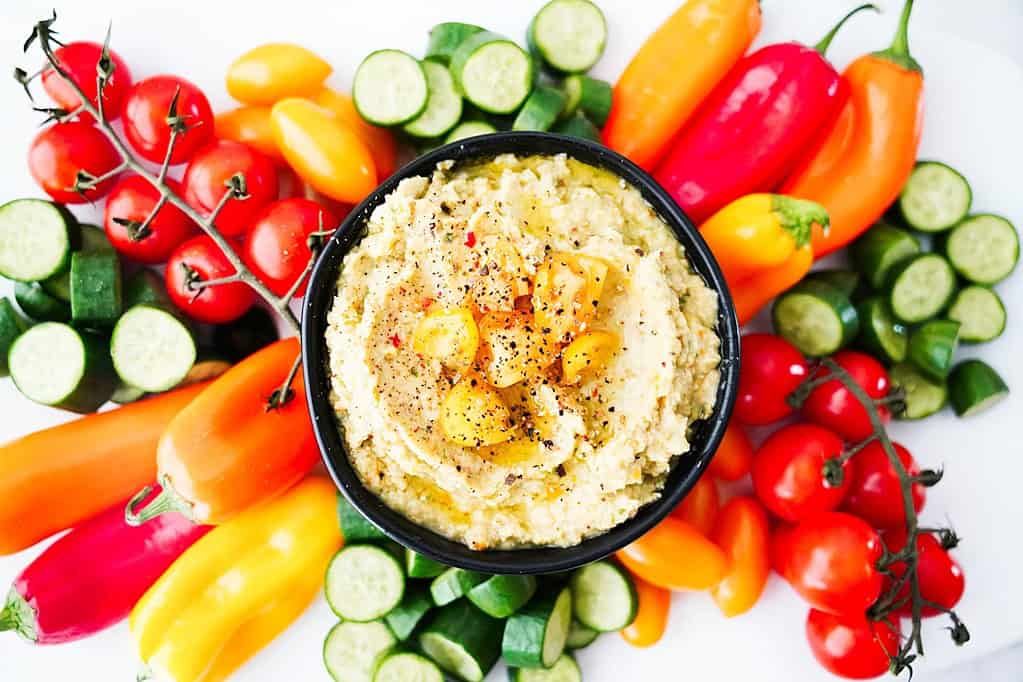Cucumbers, pepers, and tomatoes around golden tomato hummus dip