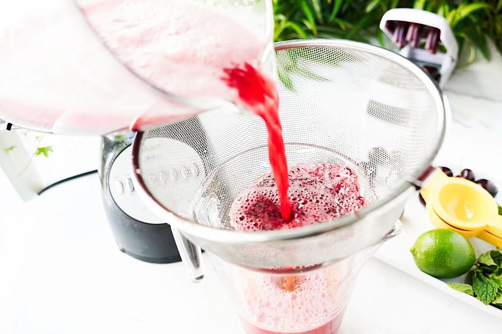 Cherry and lime agua fresca poured through a sieve, over a pitcher