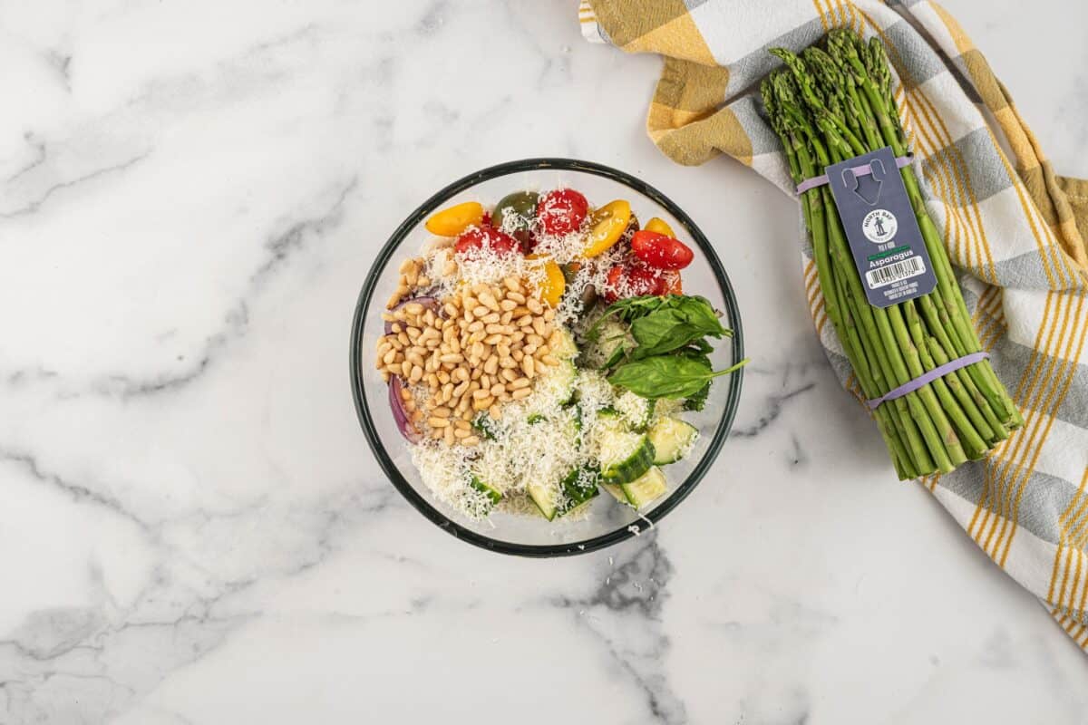 Grilled asparagus and onions, pine nuts, cucumbers, tomatoes, and parmesan cheese in a serving bowl before mixing.
